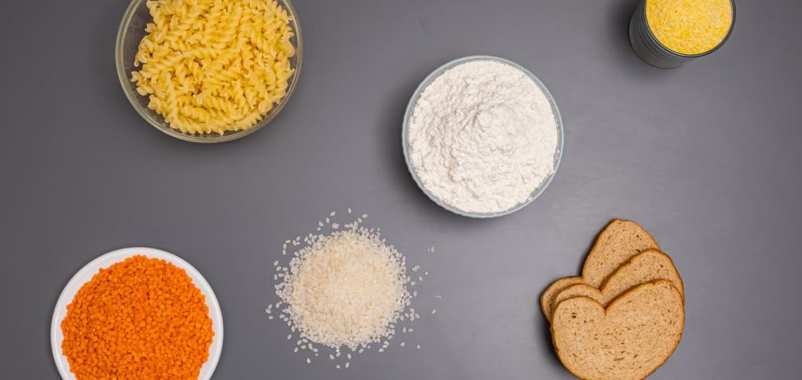 An assortment of dry ingredients including pasta, rice, and bread displayed on a grey background.